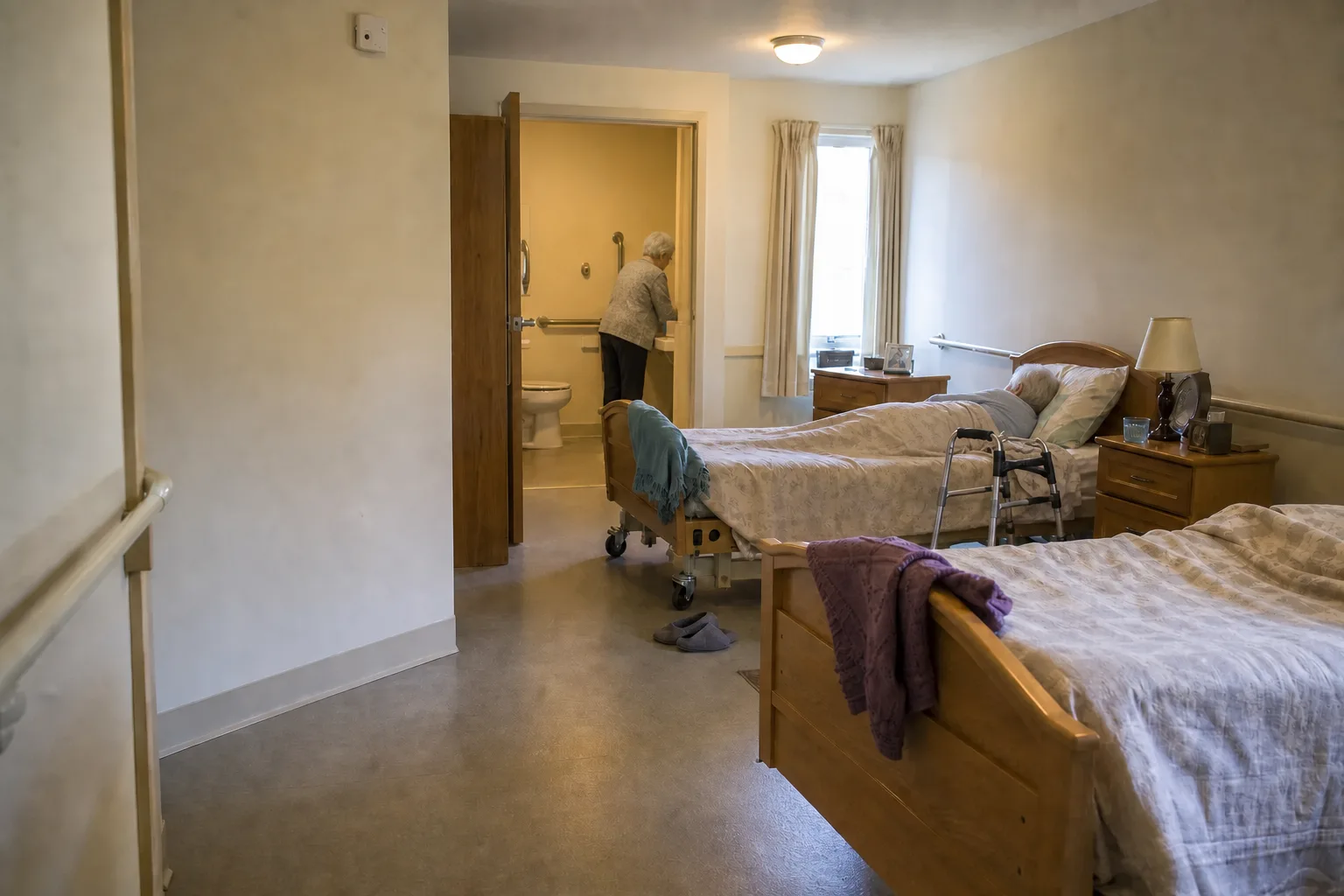 One resident resting in bed while another uses the accessible bathroom in an ordinary shared nursing-home room
